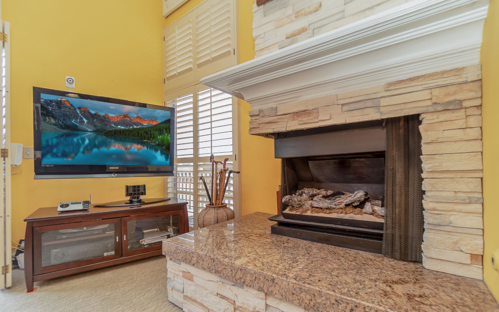 Living room with a stone fireplace, granite hearth, and television beside shuttered windows.