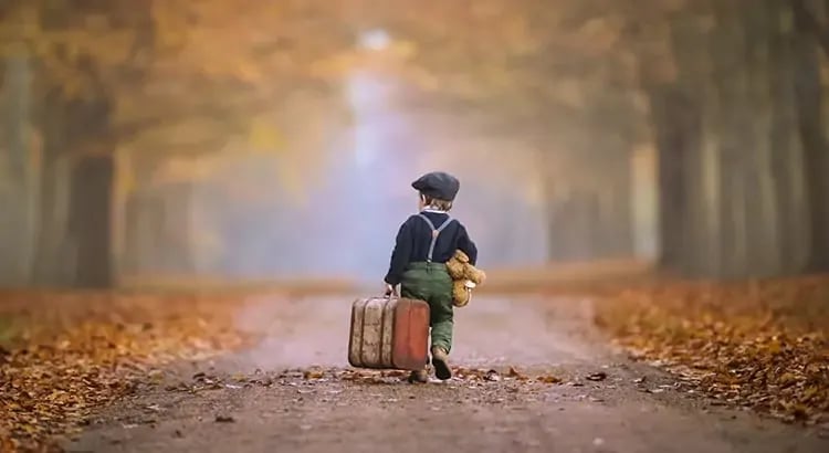 A young child carrying a suitcase in a picturesque autumn setting. The child is wearing a hat and coat, and the background features a path lined with colorful autumn leaves.