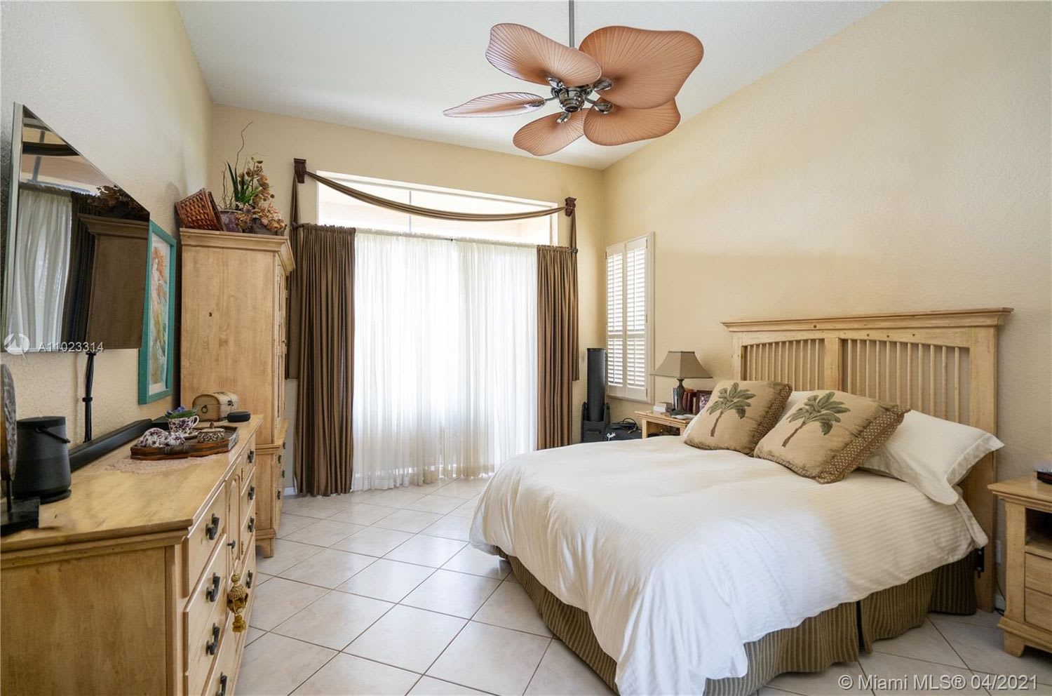 Furnished bedroom with a large bed, matching wood furniture, tiled floors, and a distinctive wood-tone ceiling fan.