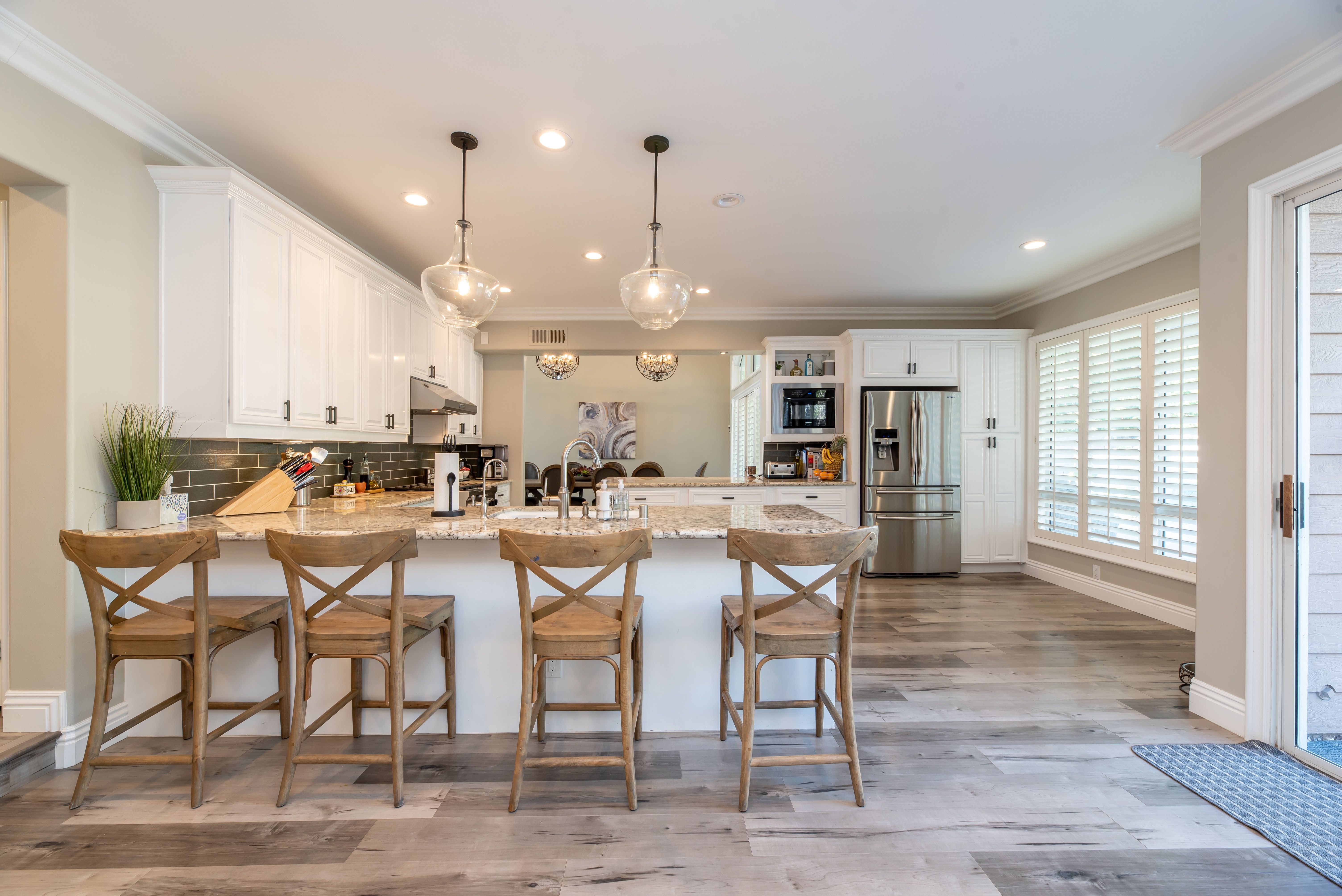 Bright kitchen with a marble-patterned island, four wooden stools, white cabinets, and plantation-shuttered windows.