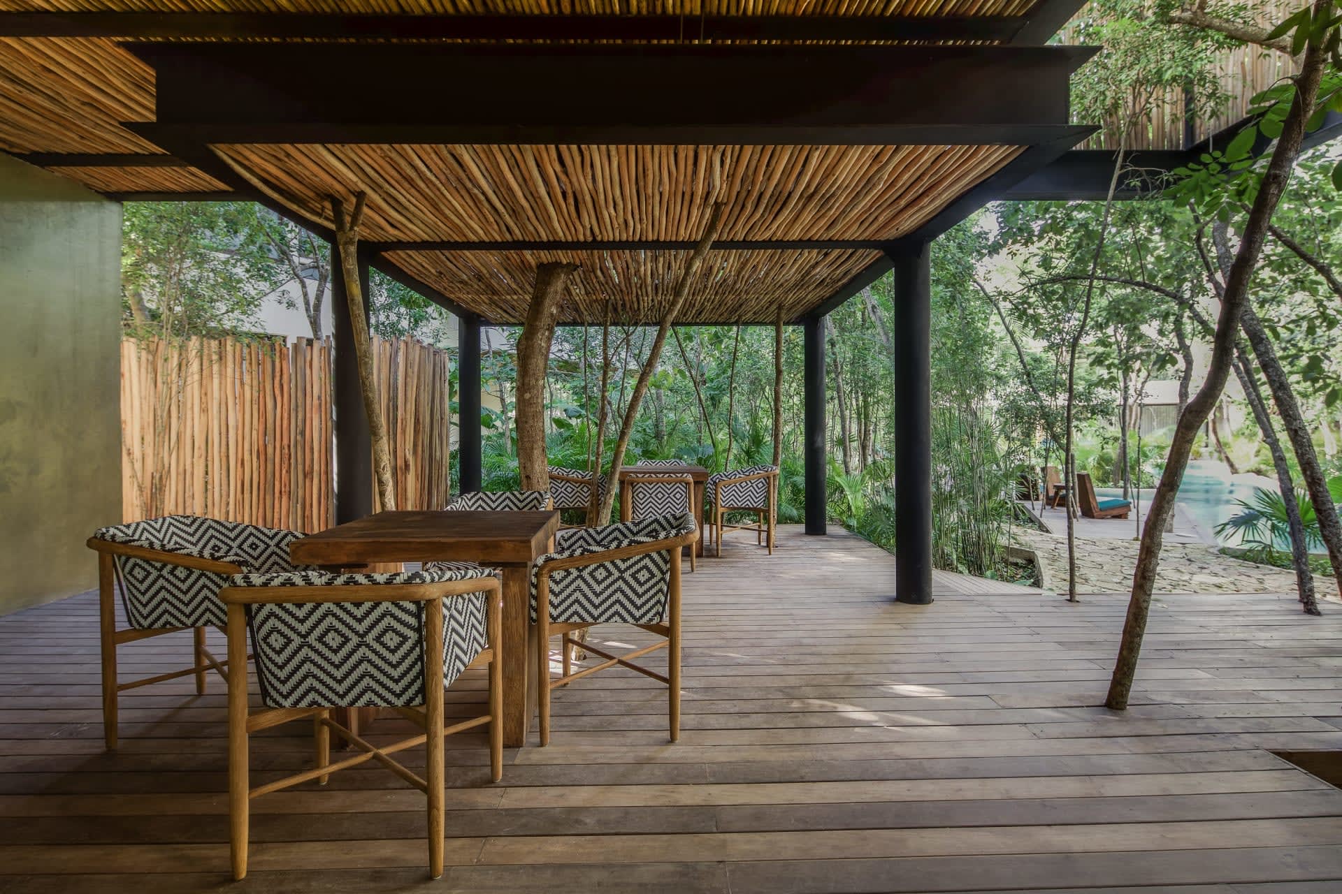 Covered outdoor dining patio with patterned chairs, a bamboo canopy, and a pool amid tropical greenery.