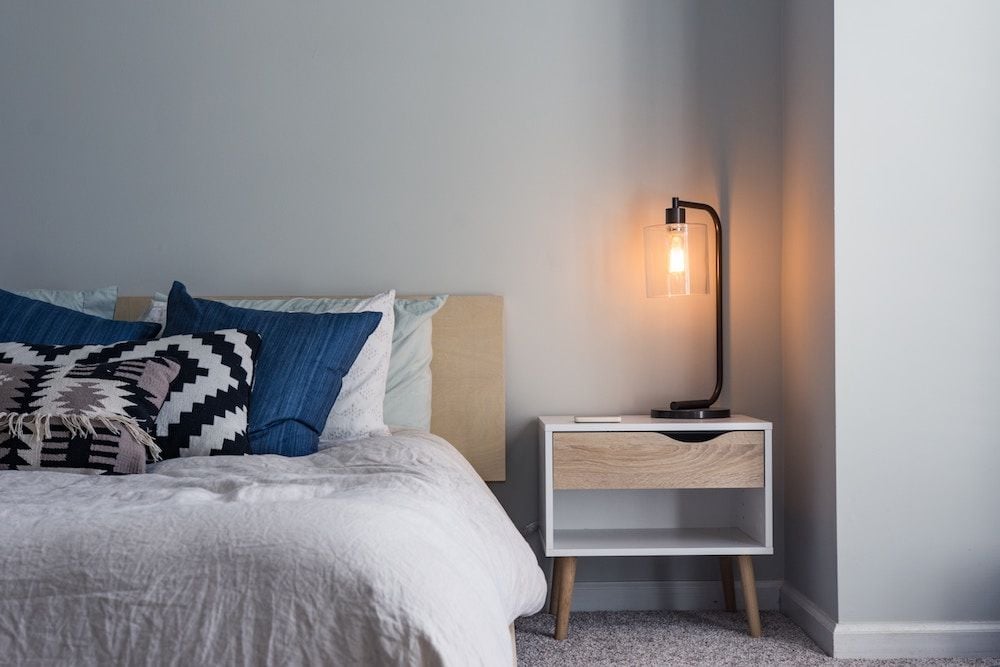Bedroom with blue and patterned pillows beside a wood-and-white nightstand and glowing lamp.