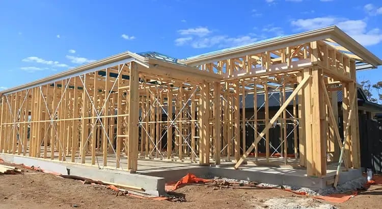 A construction site with the wooden frame of a house being built. The structure is in the early stages of construction, with a clear blue sky in the background.