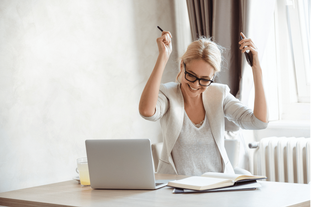 A smiling woman celebrates at her desk, raising two pens beside an open laptop and notebook.