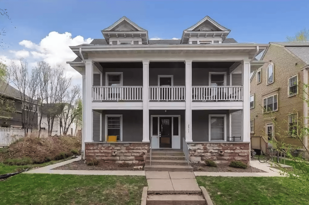 Two-story gray house with white-trimmed porches, stone foundation, and front walkway.