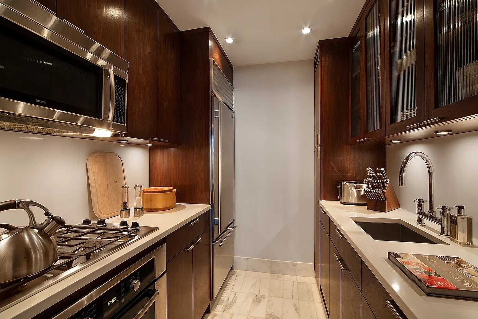 Galley kitchen with dark wood cabinets, stainless steel appliances, and white marble flooring.