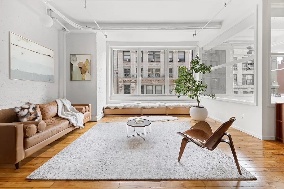 Staged living room with large city-facing windows, hardwood floors, and a tan leather sofa.