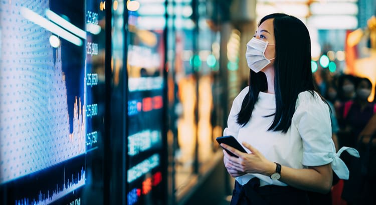 A woman wearing a protective face mask, standing in front of a large, brightly lit electronic display.