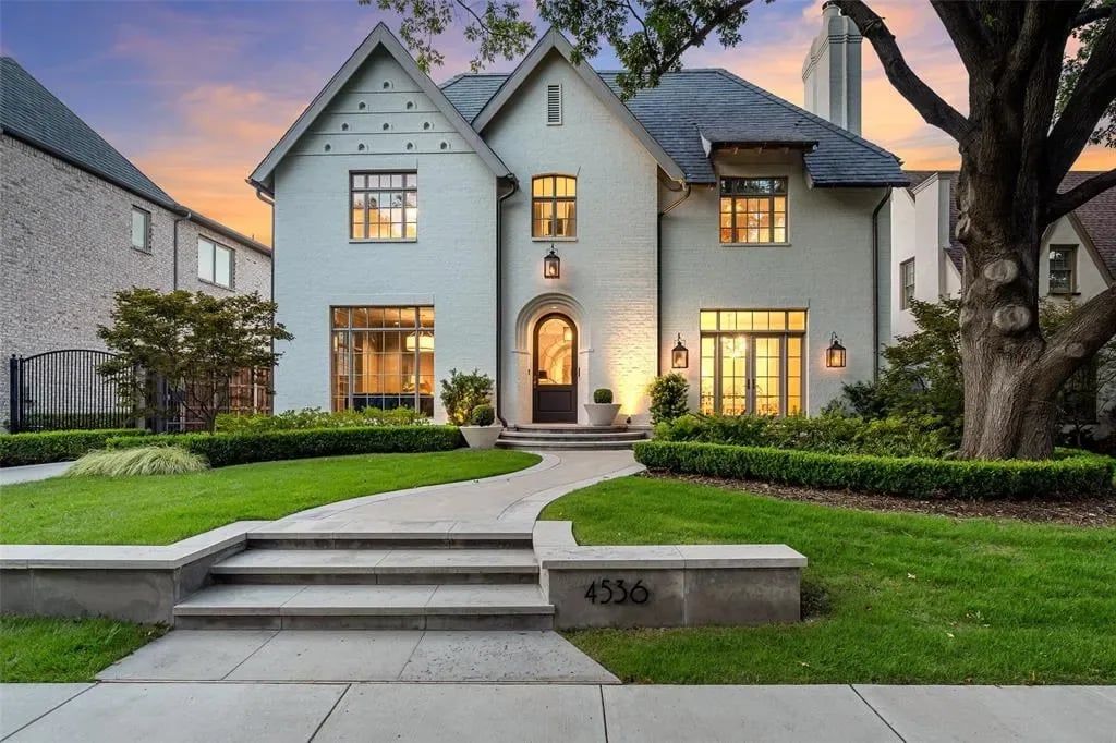 A two-story white house with a shingled roof, small porch, walkway, two-car garage, hedges, and trees on the front lawn.