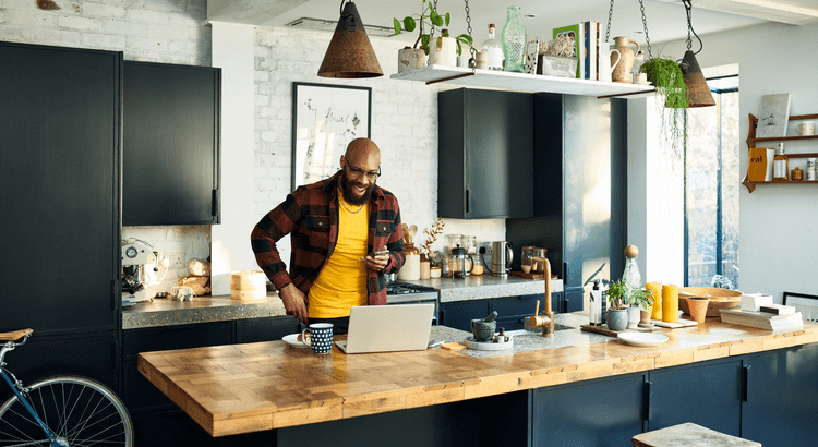 A man in a yellow shirt stands in a kitchen, focused on his laptop, surrounded by modern appliances and a bright atmosphere.