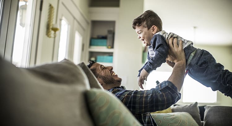 A man playing with a child in a living room. The man is lying on a couch, lifting the child above him, creating a playful and joyful scene.