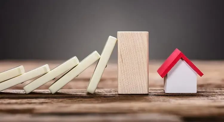 Wooden dominoes falling next to a small house model, symbolizing the concept of cause and effect in real estate or financial investments.