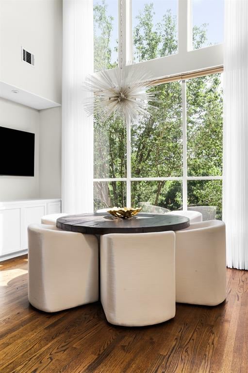Dining area with a round table, cream chairs, tall windows, and leafy trees outside.