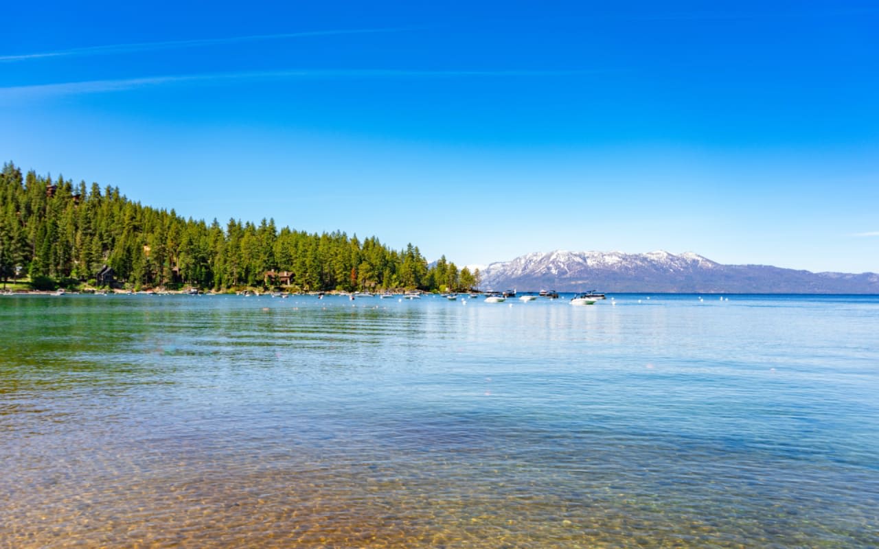 Lake Tahoe scene with clear blue skies, snow-capped mountains, and calm, crystal-clear water.