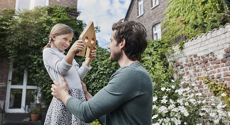 A man and a young girl are outdoors, possibly in a garden or backyard, with greenery and a brick wall in the background. The man is lifting the girl, who is smiling and holding a small, wooden birdhouse.