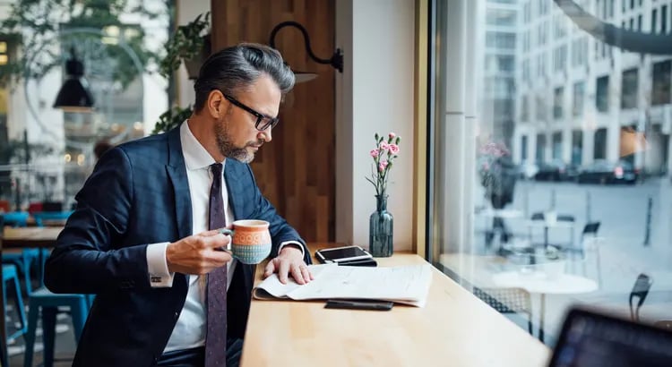 A person in a business suit sitting at a table by a large window, drinking coffee and looking at a tablet or smartphone. 