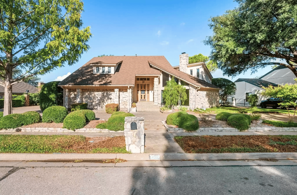 Stone-fronted home with a brown shingle roof, landscaped shrubs, and a central walkway.