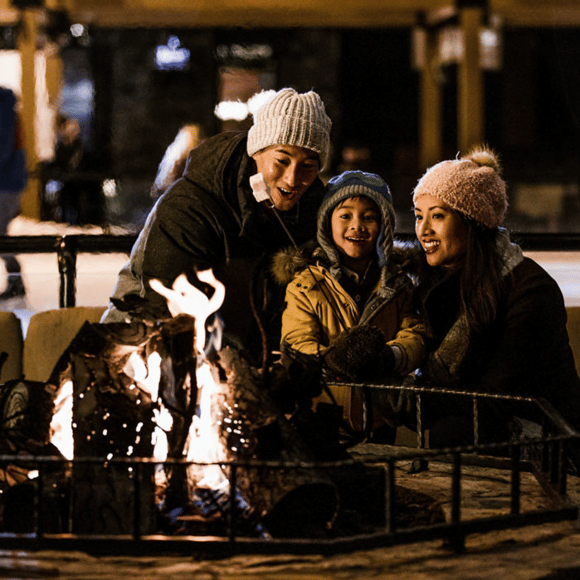 Family in winter hats gathers around an outdoor fire pit at night.