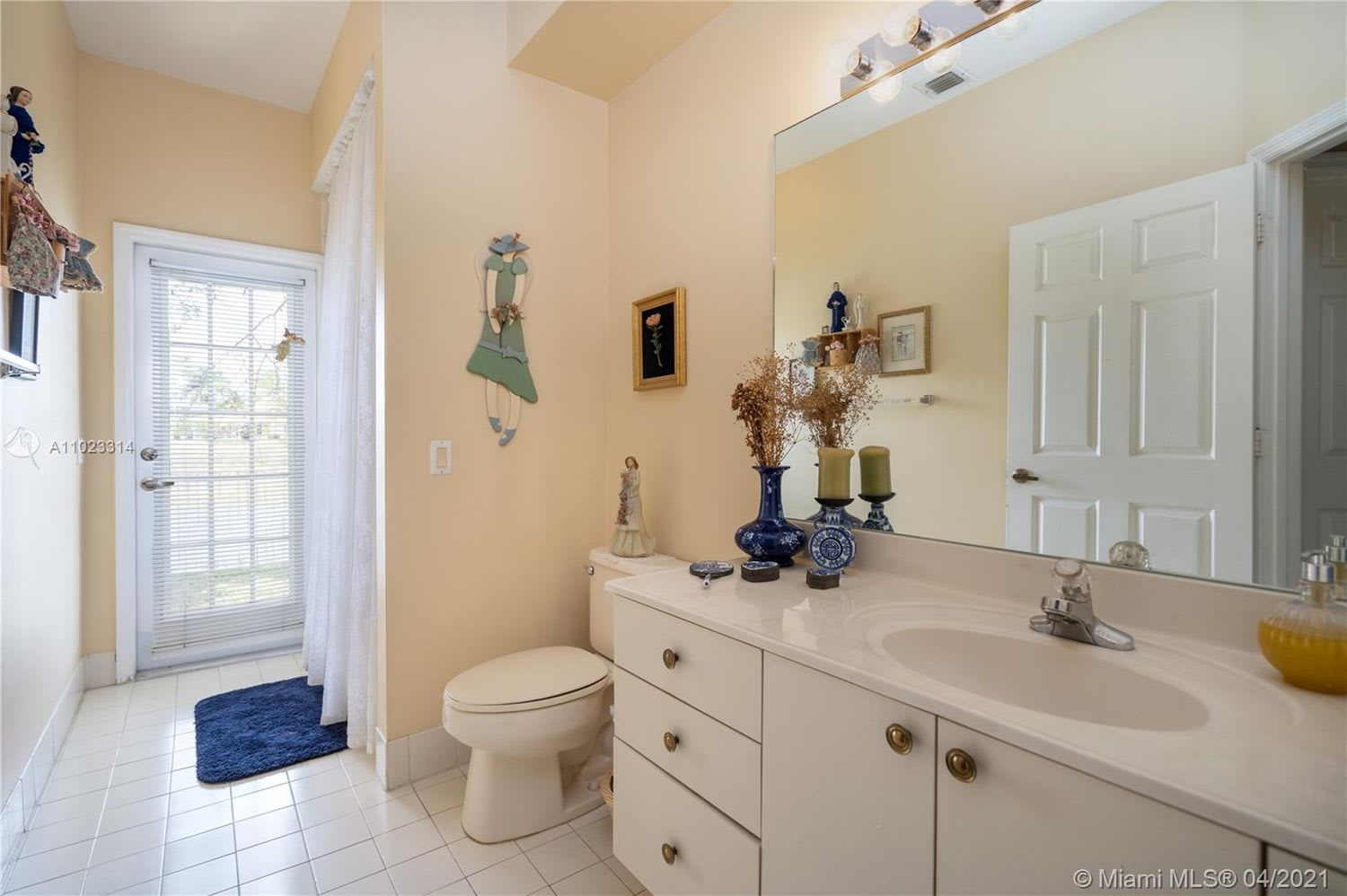 Bathroom with a long white vanity, large mirror, toilet, and glass-paneled door with blinds.