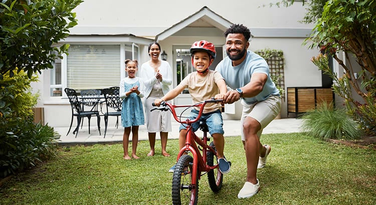 A family scene with parents helping their child ride a bicycle. They are outdoors in a yard with green grass and a house in the background.