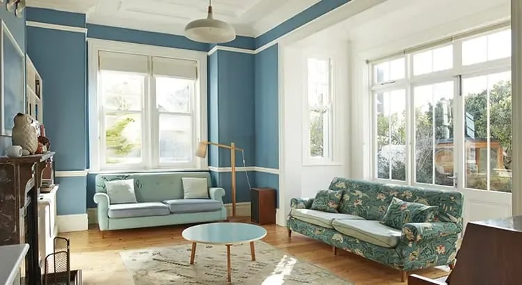 A brightly lit living room with blue walls and large windows. The room features a cozy seating area with a sofa, chairs, and a coffee table.