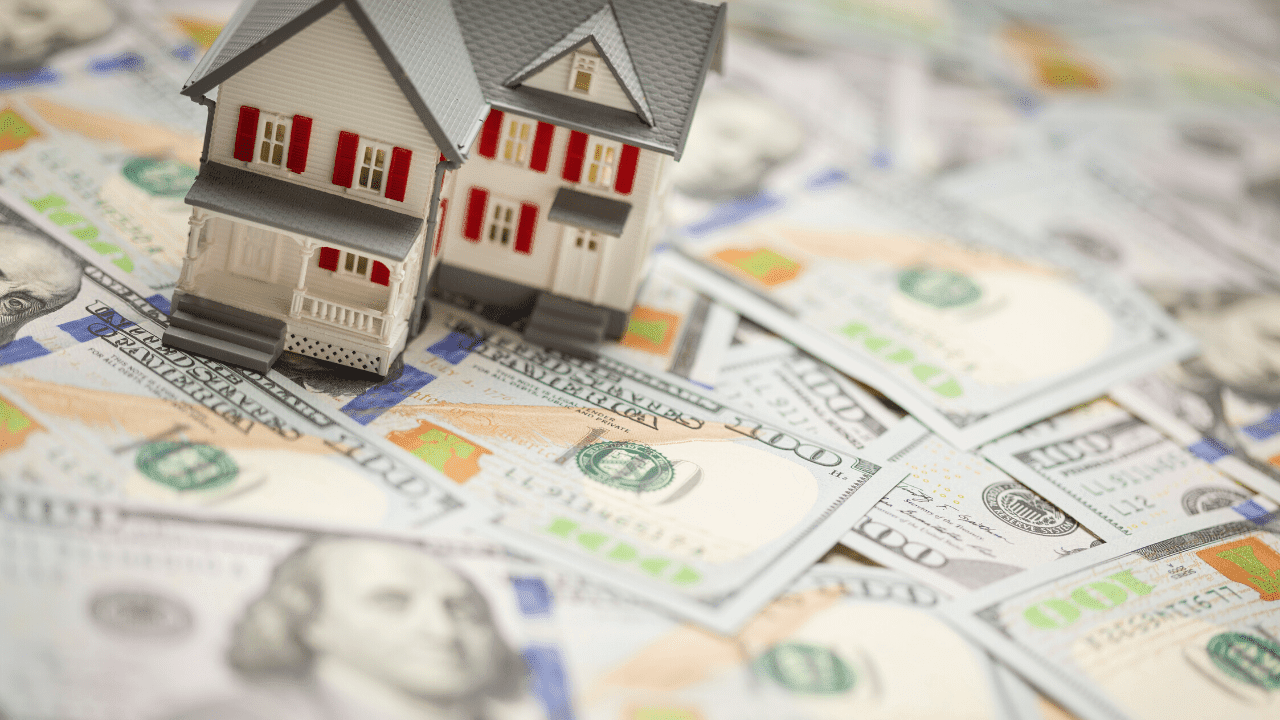 A small model house positioned on top of a spread of U.S. hundred-dollar bills. 