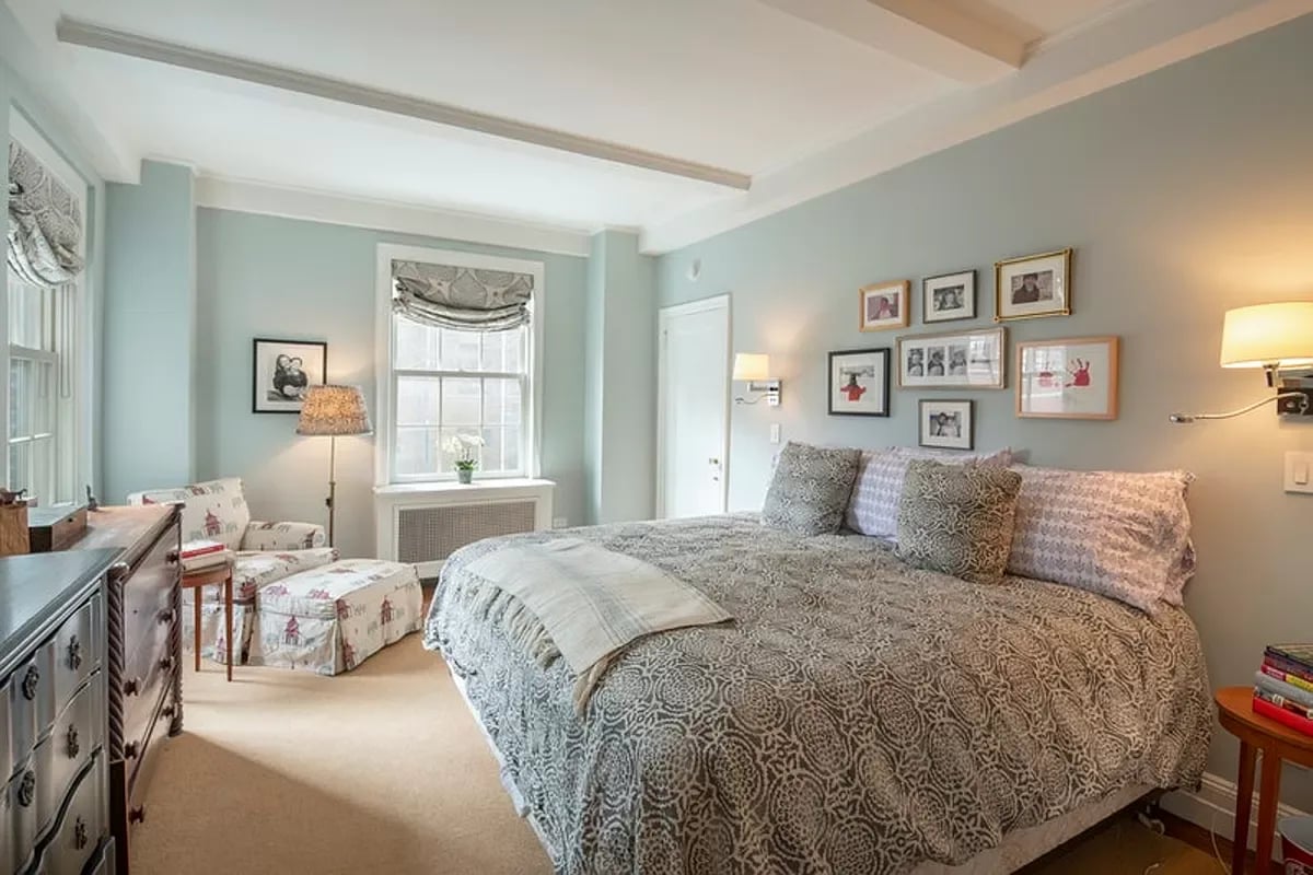 Bedroom with a large patterned bed, pale blue walls, and a small sitting area beside the windows.