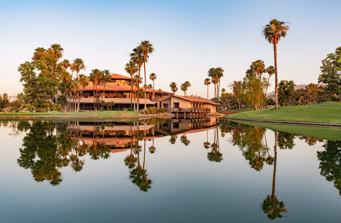 a large, modern clubhouse with a red tile roof surrounded by palm trees and a reflecting pond