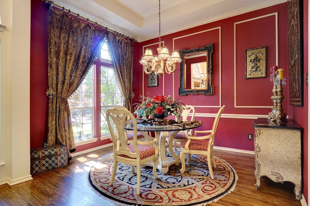 Dining room with red paneled walls, ornate chairs, a glass-top table, and tall curtained windows.