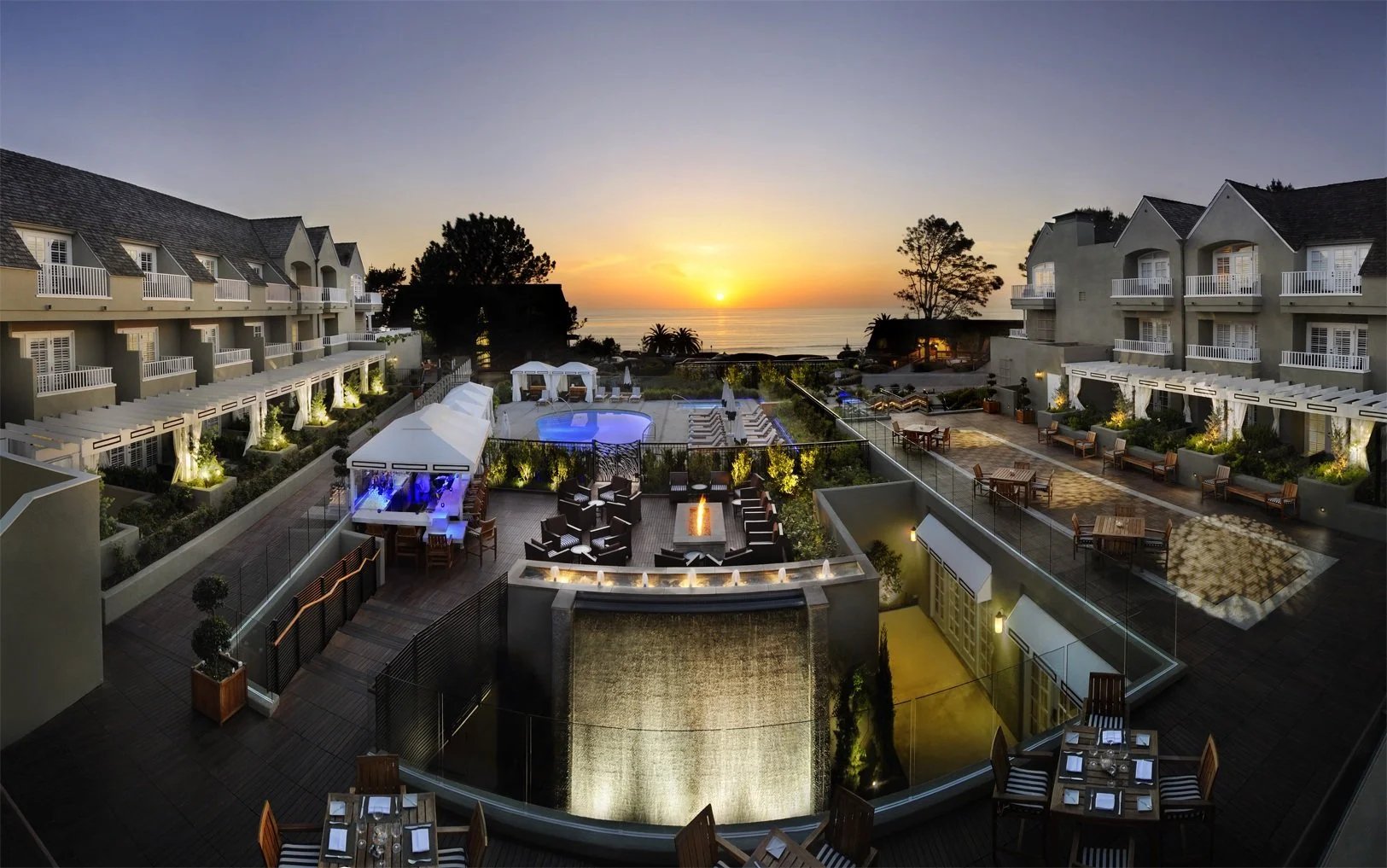 Residential courtyard with a pool, waterfall, and ocean sunset beyond the surrounding buildings.