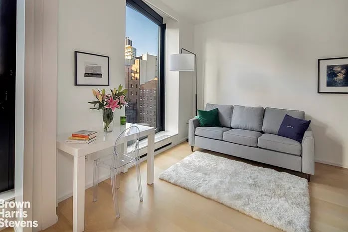 Bright living room with a gray sofa, white shag rug, and large window overlooking city buildings.