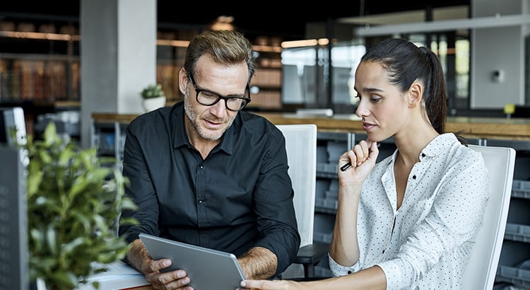 Two people, a man and a woman, are seated at a table, engaged in a discussion. The man is wearing glasses and a black shirt, and the woman has her hair tied back and is wearing a white shirt. They appear to be in a professional setting, possibly an office or a café.