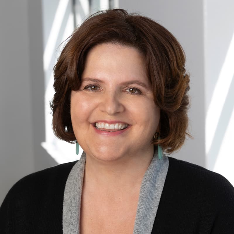 Jennifer Burden, Founder and Associate Broker of Legacy Real Estate, and top 1% San Francisco realtor serving Bernal, Bayview, and beyond. She has short brown hair, is wearing a black top with a gray collar, and is smiling at the camera. The background features a light-colored wall with geometric patterns created by shadows and light.