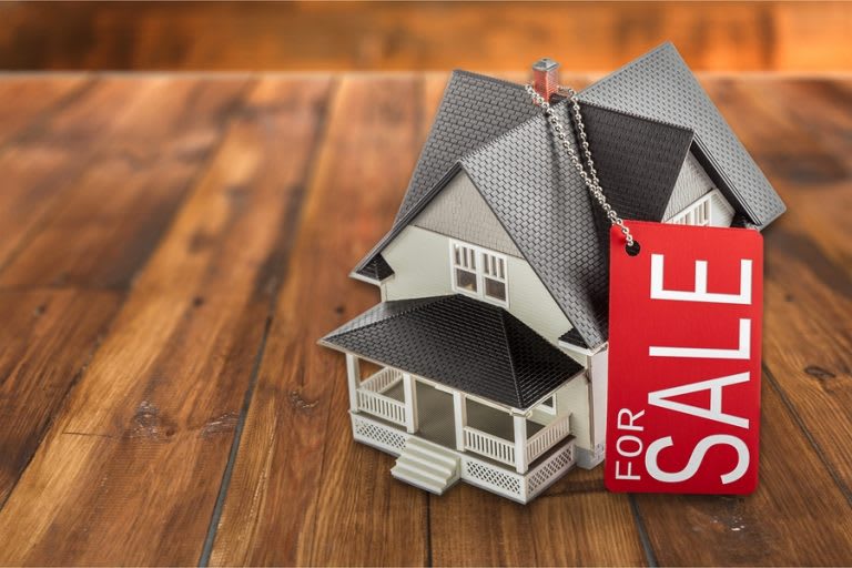 Miniature house with a red FOR SALE tag on a wooden surface.