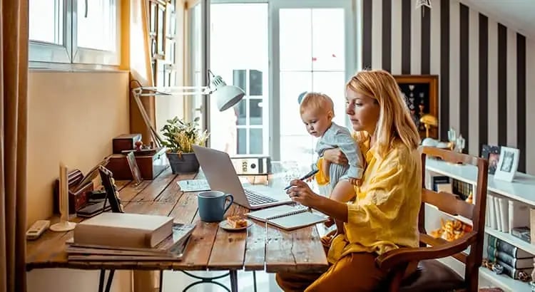 A woman working at a desk with a laptop while a child sits beside her, engaged in their own activities. The setting is a home office or study area, suggesting a work-from-home environment.