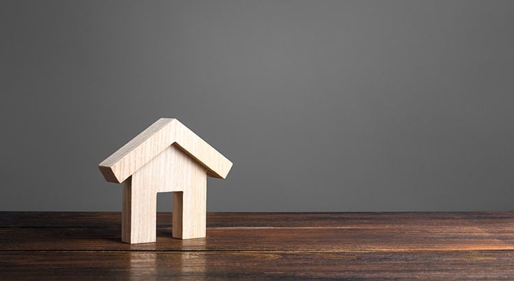 A small wooden house model on a table against a grey background. This minimalist image symbolizes homeownership, real estate, or investment.