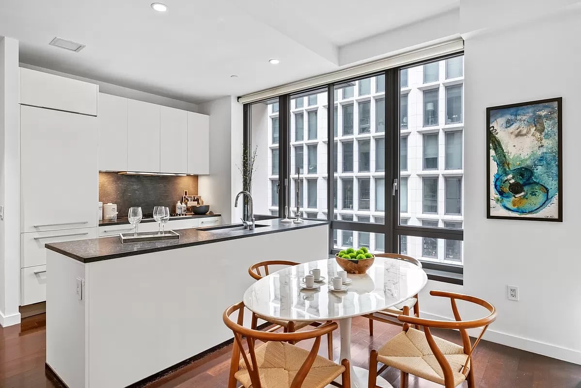Modern kitchen and dining area with a dark island, oval table, and large windows overlooking a neighboring building.