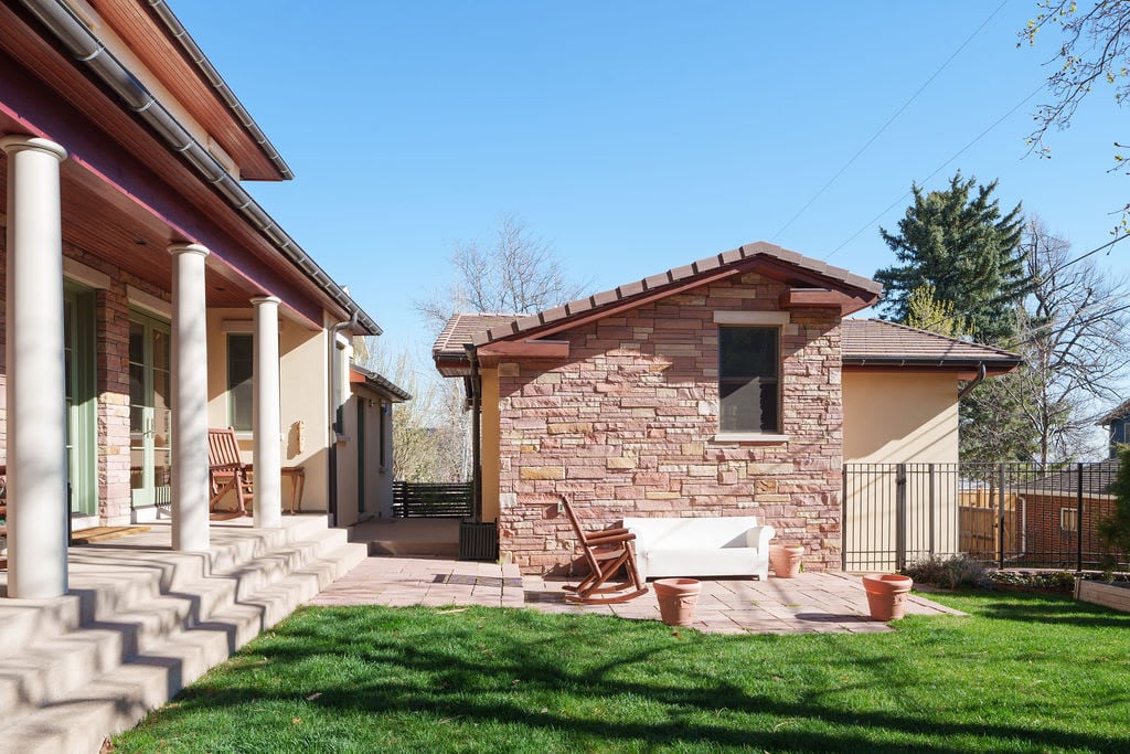 Modern home exterior with white columns and stone facade, featuring adjacent guesthouse, patio space, and manicured lawn.