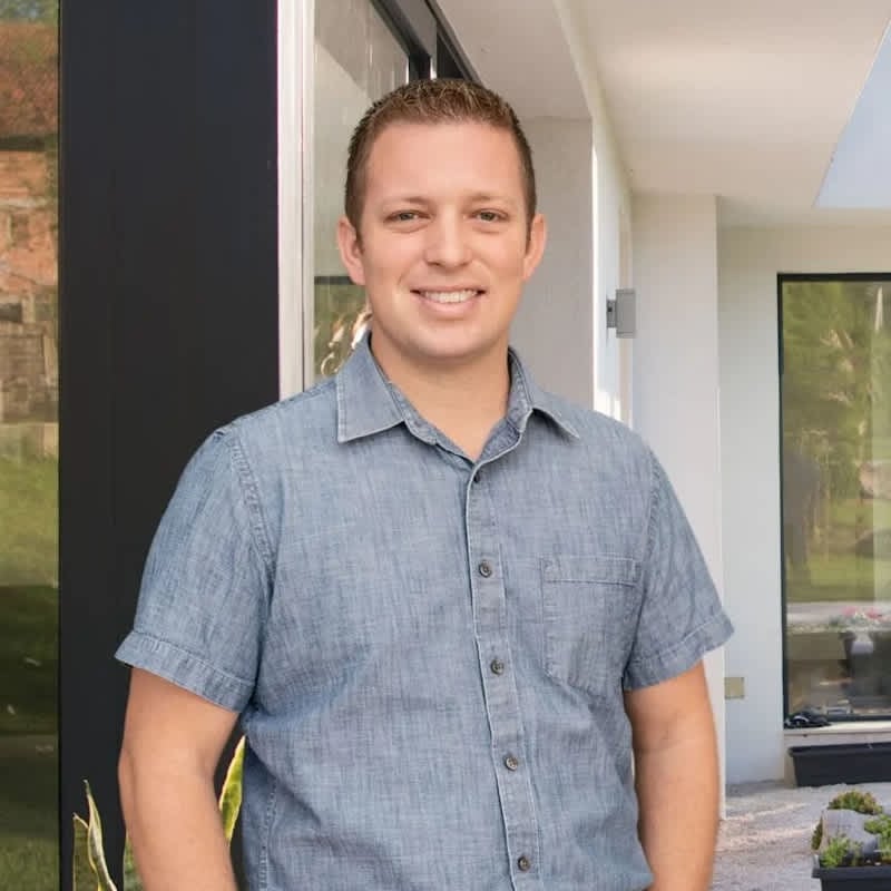 Smiling man in a short-sleeved, light blue button-down shirt standing in front of a modern house with large windows and a landscaped entryway.