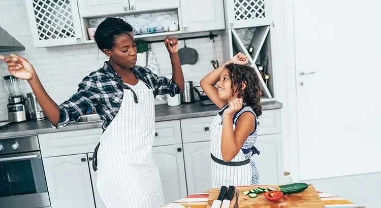 A woman and a young girl in a kitchen, wearing aprons and enjoying a playful moment while preparing food. They are both smiling, indicating a joyful and bonding activity.