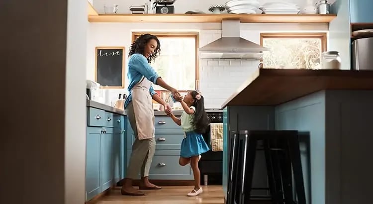 A cozy kitchen scene with a parent, a mother, cooking while a child, likely a daughter, is standing nearby. The kitchen has modern appliances, wooden cabinets, and a warm atmosphere.