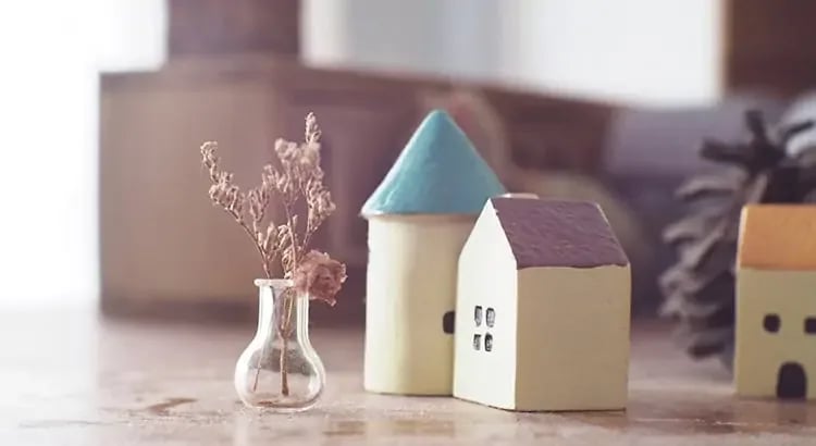 A small model house with a blue roof next to a vase with dried flowers, set on a table. The scene has a minimalist and cozy feel.