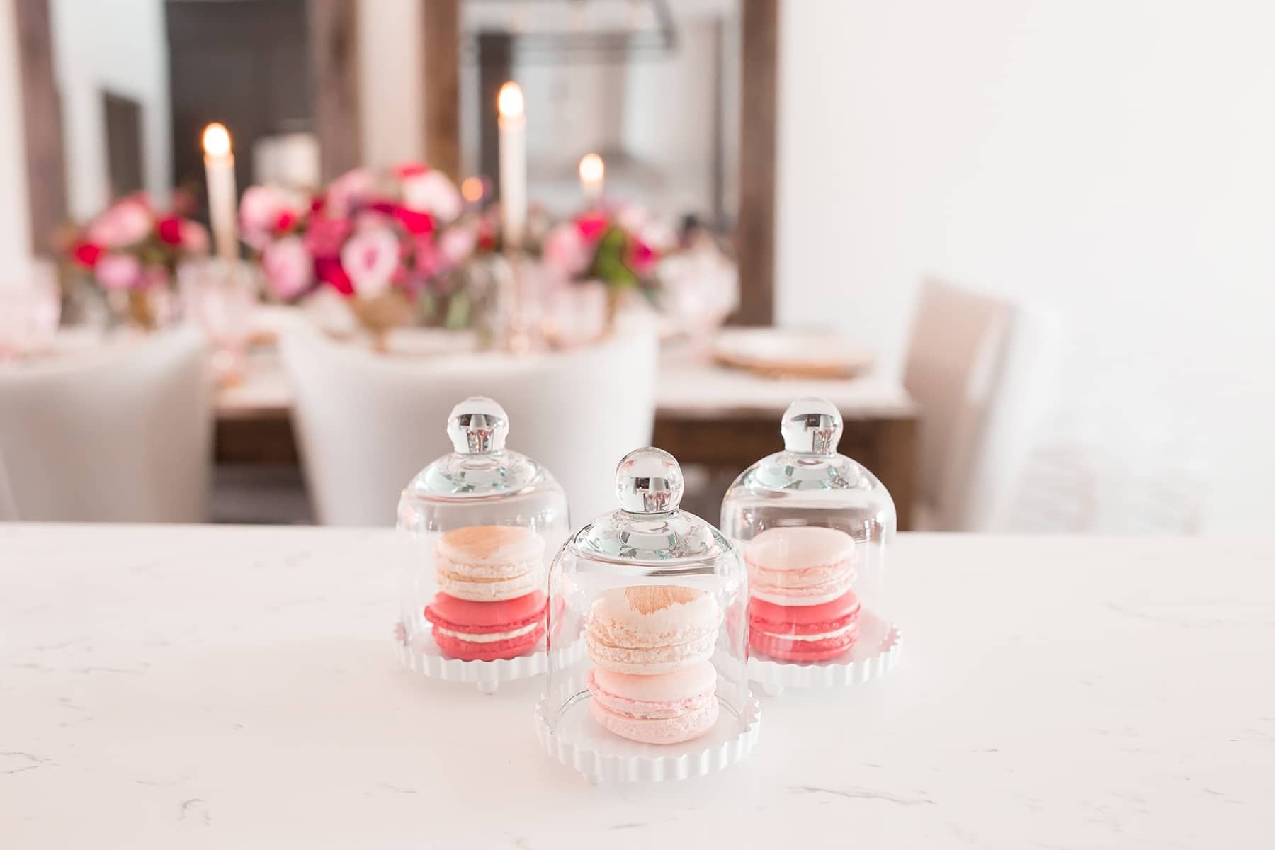 Pink and red macarons under glass domes on a marble counter, with candlelight and pink flowers behind them.