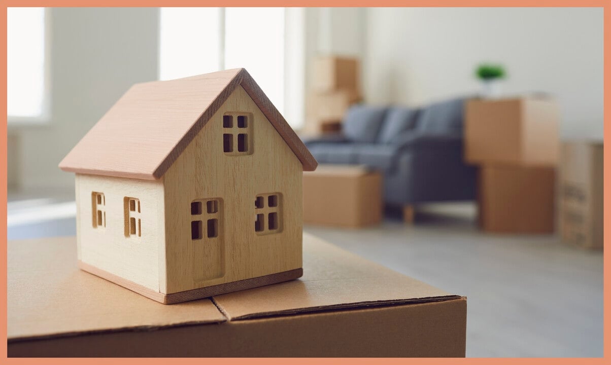 Wooden model house on a moving box, with packed boxes and a sofa in the background.