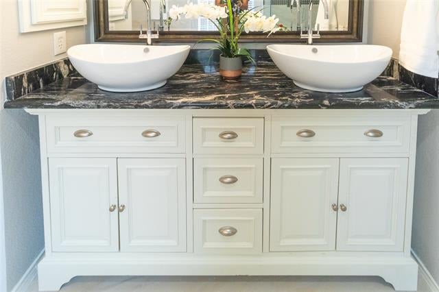 Bathroom vanity with twin vessel sinks, dark marble countertop, white cabinetry, and an orchid.