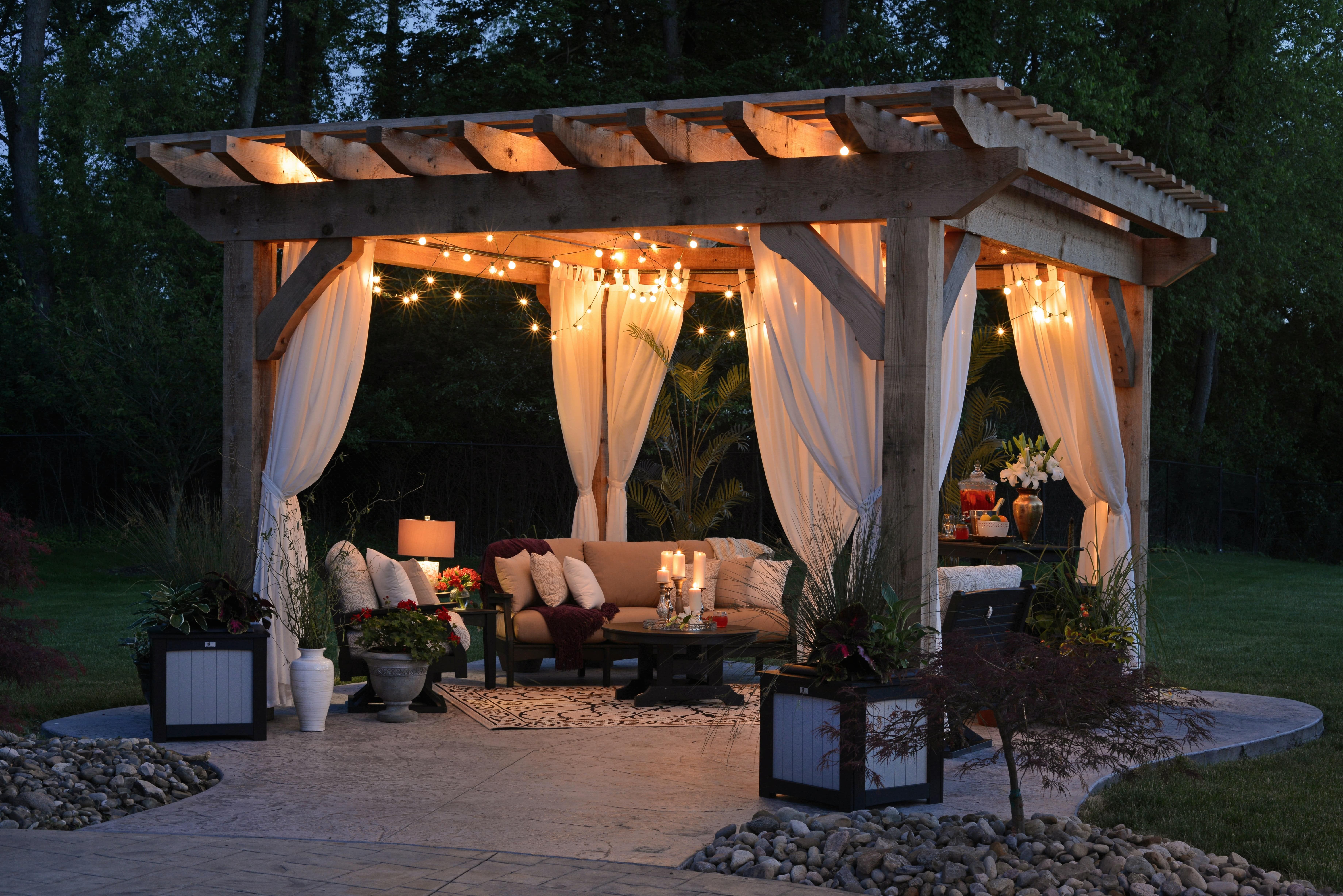 Backyard patio under a wooden pergola, with curtains, string lights, and comfortable seating.
