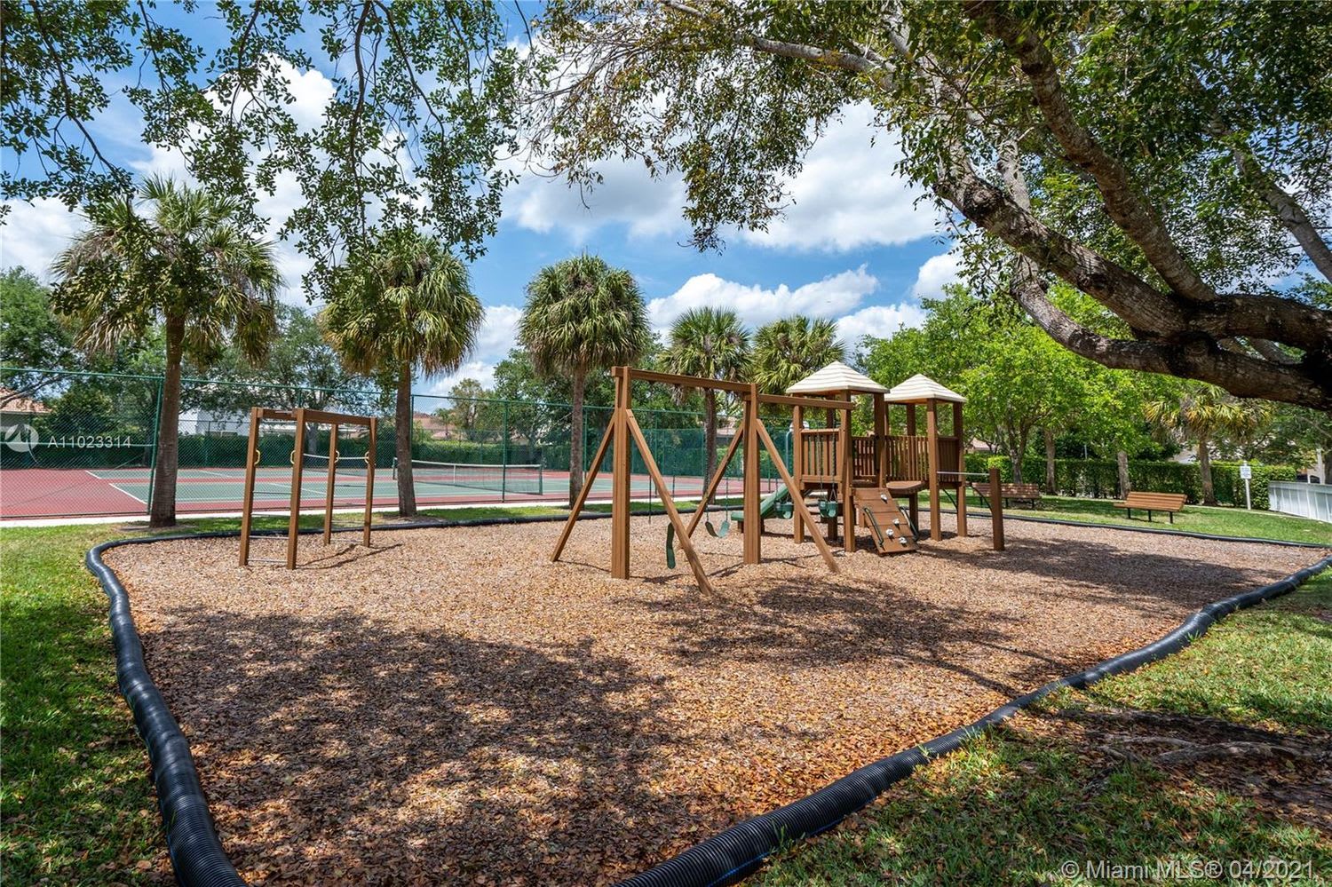 Community playground with wooden play equipment and swings beside a fenced tennis court.