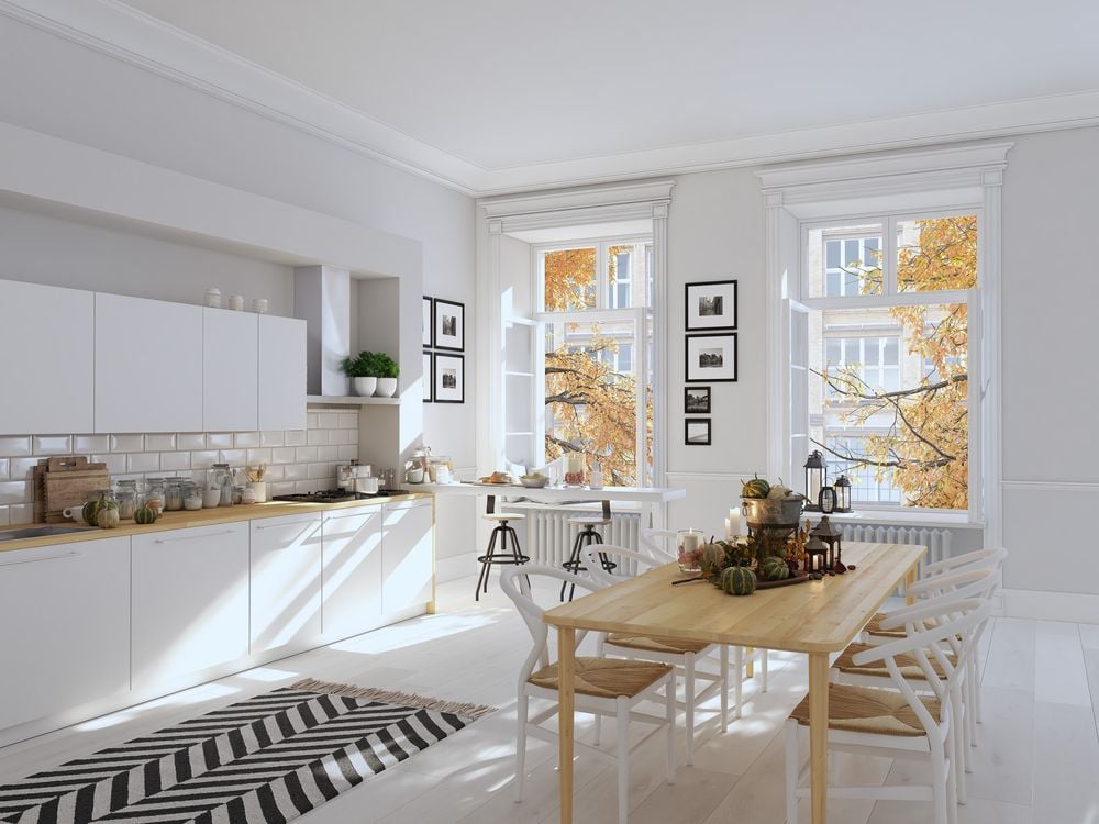 Bright white kitchen and dining area with autumn decorations on a long wooden table and fall trees outside.