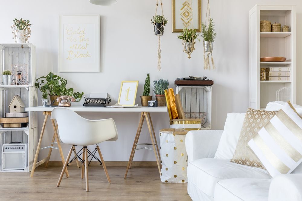 Bright living room workspace with a trestle-leg desk, plants, gold accents, and a white sofa.