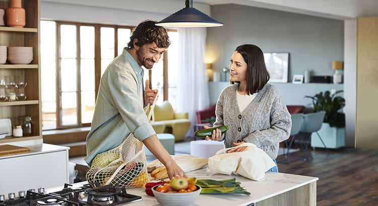 A scene of a kitchen with two people preparing food. They are standing at a kitchen island with various fruits and vegetables. The kitchen has a modern design with wooden cabinets and a window in the background.